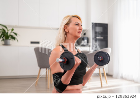 Focused blonde woman sitting on yoga mat lifting dumbbell for bicep training during home fitness session in modern living room Focused blonde woman sitting on yoga mat lifting dumbbell for bicep training during home fitness session in modern living room 129862924