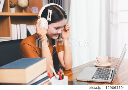 Asian girl doing homework and studying at home. woman using laptop at desk write notes watching webinar, study online, look at learn web class or having virtual call meeting remote working from home. 129863394
