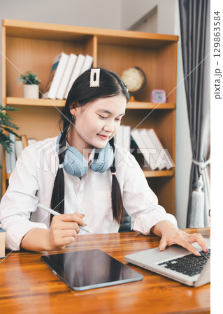 Asian girl student wear headphones learning online watching webinar class looking at laptop computer eLearning remote lesson making notes or video calling virtual conference meeting teacher at home. 129863584