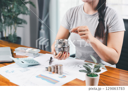 Young Asian woman making stack of coin. invest save finance concept, saving money, investment. dollar bills on table with pile of coins and banks calculator, money to save and invest to make income. 129863783