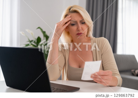 Blonde woman in beige outfit sitting at home, looking upset at a bill and struggling with financial difficulties, economic stress concept Blonde woman in beige outfit sitting at home, looking upset at a bill and struggling with financial difficulties, economic stress concept 129863792