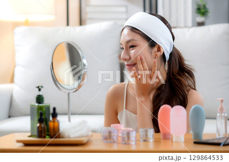 Beauty young asian woman applying cream on facial for wrinkles while looking mirror in living room. Beauty young asian woman applying cream on facial for wrinkles while looking mirror in living room. 129864533