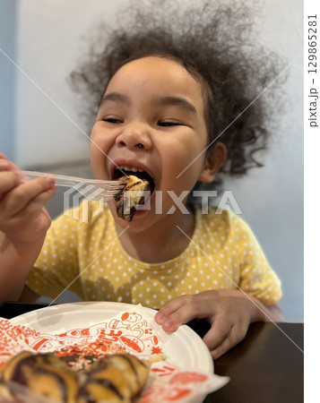 Cute little Asian girl eating a piece of chocolate donut in cafe with fork 129865281