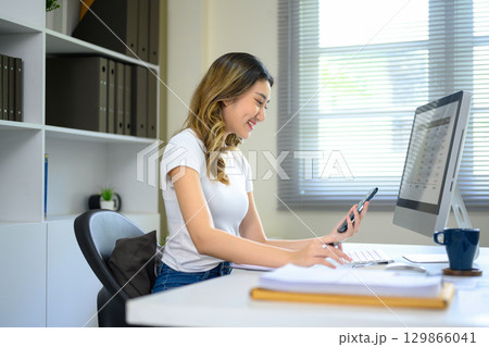 Happy female worker analyzing financial charts on paper and using mobile phone at computer workstation 129866041