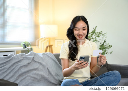 Smiling woman holding a credit card and smartphone while making an online payment at home 129866071