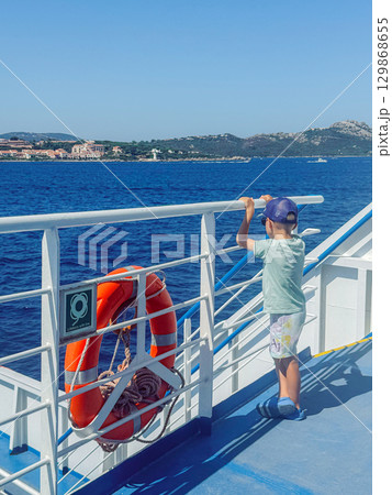Young boy looks out at the sea from the ferry deck, holding the railing for a safe view. The life buoy signifies safety aboard during the journey across calm waters 129868655