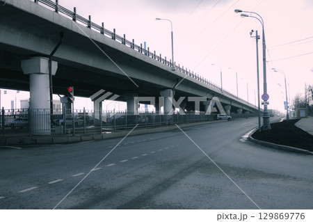 Modern city transport interchange with a high overpass bridge, supports and street lamps. Empty asphalt road goes into the distance under a gray sky. Cool atmosphere, urban, with a feeling of silence Modern city transport interchange with a high overpass bridge, supports and street lamps. Empty asphalt road goes into the distance under a gray sky. Cool atmosphere, urban, with a feeling of silence 129869776