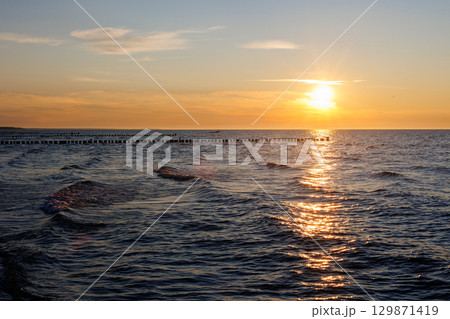 Scenic warm sea sunset view golden sunset over Baltic Sea reflects across rippling waves, creating a glowing path of light. Wooden breakwaters stretch into horizon calm rhythm to tranquil seascape 129871419