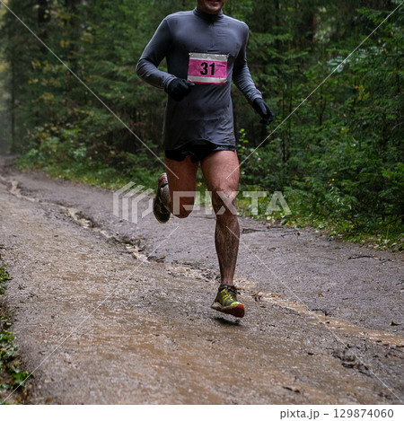Runner in numbered bib running muddy forest trail during marathon race 129874060