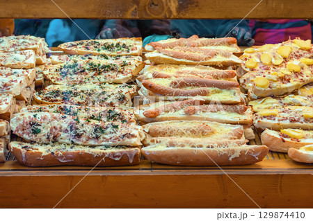 Freshly baked baguettes topped with sausages, cheese, herbs, and potatoes at food stall. Concept of cheesy sausages, French hotdog, comfort fast food, festive street meals Freshly baked baguettes topped with sausages, cheese, herbs, and potatoes at food stall. Concept of cheesy sausages, French hotdog, comfort fast food, festive street meals 129874410