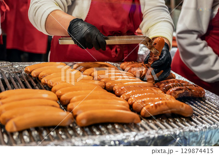 Grilled sausages at Christmas food stall. Vendor in gloves grills sausages on open flame at busy Christmas food stall during winter market. Concept of street food, festive culinary tradition Grilled sausages at Christmas food stall. Vendor in gloves grills sausages on open flame at busy Christmas food stall during winter market. Concept of street food, festive culinary tradition 129874415