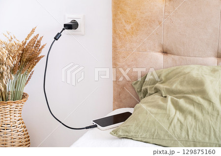Phone charging cable connected to wall socket next to bed. 129875160