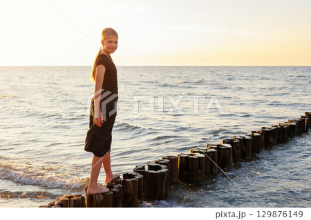 Cute adorable blond teenager happy kid girl walking by wooden breakwater posts at Baltic sea scenic warm summer sunset light. Balancing reflects challenges, choices and uncertainties of growing up Cute adorable blond teenager happy kid girl walking by wooden breakwater posts at Baltic sea scenic warm summer sunset light. Balancing reflects challenges, choices and uncertainties of growing up 129876149