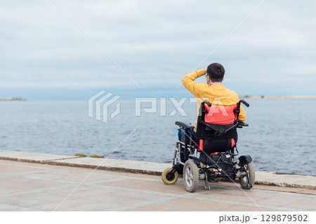 Rear view of disabled Caucasian woman sitting with arms up on wheelchair at beach. Rear view of disabled Caucasian woman sitting with arms up on wheelchair at beach. 129879502
