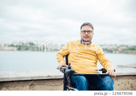Smiling young disabled man use phone on a wheelchair in a park at sunlight outdoors. 129879506