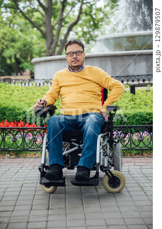 Smiling young disabled man use phone on a wheelchair in a park at sunlight outdoors. 129879507