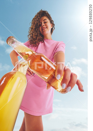 Woman in pink dress clicking beer bottle with confident smile outdoors 129880250