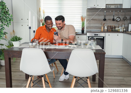 Interracial couple looking at smartphone while having breakfast in the kitchen Interracial couple looking at smartphone while having breakfast in the kitchen 129881336