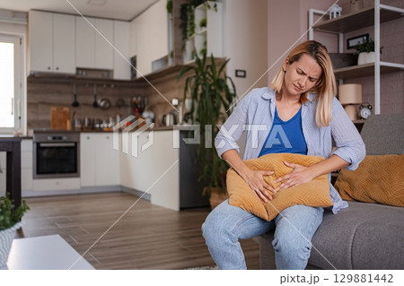 Woman suffering from strong abdominal pain while sitting on sofa at home 129881442