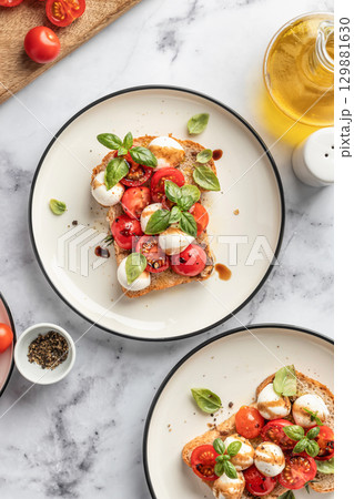 Caprese sandwich or toast with mozzarrella balls, cherry tomatoes, olive oil and fresh basil leaves top view. Healthy vegetarian breakfast, snack or lunch. White marble background Caprese sandwich or toast with mozzarrella balls, cherry tomatoes, olive oil and fresh basil leaves top view. Healthy vegetarian breakfast, snack or lunch. White marble background 129881630