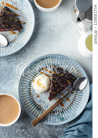 A piece of chocolate brownie cake on blue plate served with a scoop of ice cream and caramel topping with coffee, top view 129881754