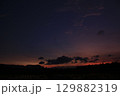 Peaceful rural scene of a rice field under a twilight sky. Dark clouds drift across a gradient of orange and purple hues, with silhouettes of trees and hills in the distance. 129882319