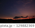 Wide view of a colorful sky transitioning from orange to purple above a flooded rice field at sunset. Dark clouds and distant mountain silhouettes create a peaceful mood. 129882369