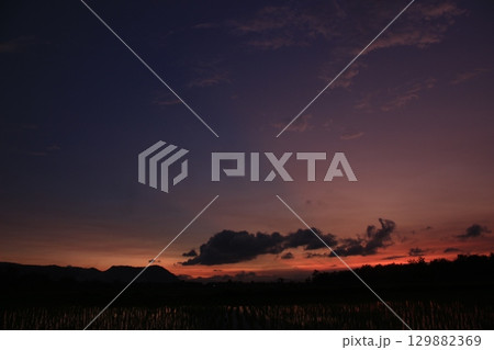 Wide view of a colorful sky transitioning from orange to purple above a flooded rice field at sunset. Dark clouds and distant mountain silhouettes create a peaceful mood. 129882369
