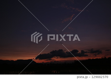 A moody twilight landscape featuring dark cloud shapes against a fading orange and purple sky. Silhouettes of distant hills and flooded rice fields evoke a calm rural scene. 129882370