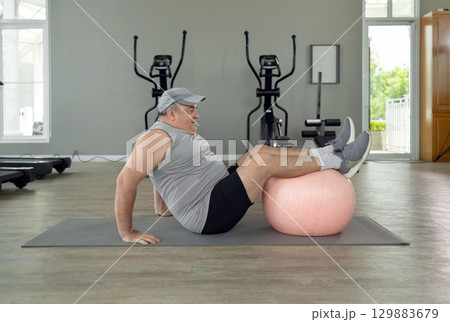 Senior chubby guy doing physical exercise with fitness ball lying on floor at gym. Side view of man having sport workout after gaining weight and getting fat. 129883679