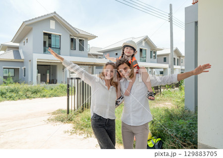 A cheerful family celebrate their new home in a developing neighborhood. The parent wear casual clothing while holding their child in a safety vest and hardhat, creating a lively atmosphere. 129883705