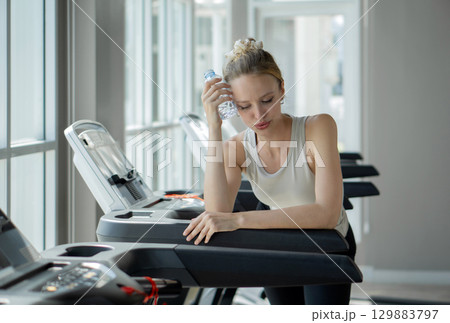 A woman take a break while exercising on a treadmill, holding a water bottle and catching her breath in a bright, contemporary gym setting with large window. 129883797