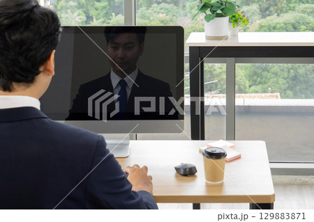 A man in formal attire participate in a video conference from his workspace. A computer screen reflect his image while he focus on the discussion. Outside, greenery add to the atmosphere. 129883871