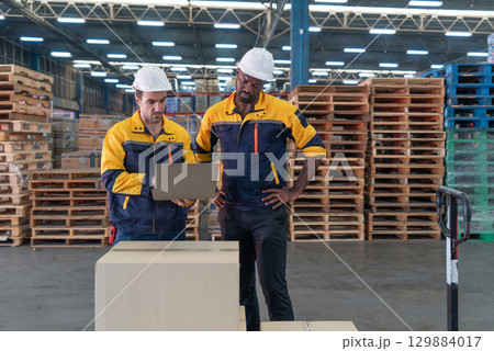 Two worker wearing safety helmet are focused on a laptop, stand in front  a large cardboard box. They are engaged in a discussion in a busy warehouse filled with wooden pallet. 129884017