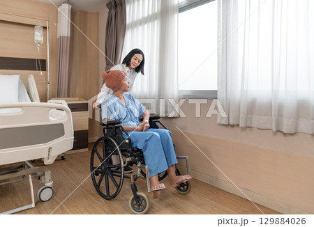 A caregiver support a chemo patient with knitted hat on her head sitting in wheelchair, they enjoy the natural light coming through the window in hospital room. The atmosphere is calm and comforting. A caregiver support a chemo patient with knitted hat on her head sitting in wheelchair, they enjoy the natural light coming through the window in hospital room. The atmosphere is calm and comforting. 129884026