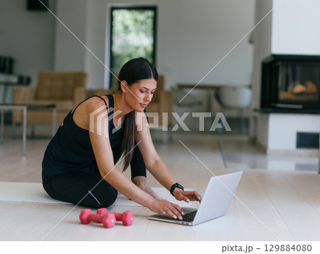 A young woman in sportswear is sitting in the living room and preparing for online training while using a laptop. 129884080