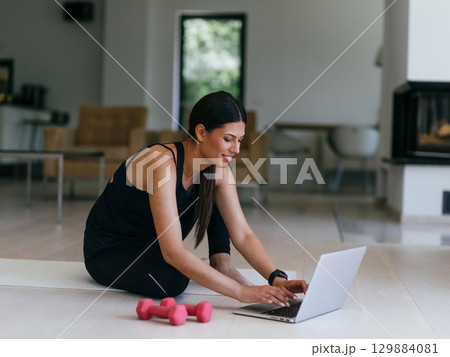 A young woman in sportswear is sitting in the living room and preparing for online training while using a laptop. 129884081