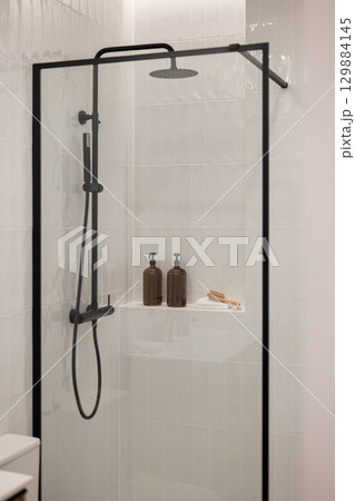 Modern shower with glass enclosure, black fixtures, and minimalist design. Two brown bottles on a shelf add a touch of elegance. 129884145