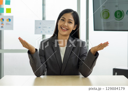 Young asian female employee in gray suit, smiling and gesturing with her hand while sit at her desk. There was a note paper and document on glass board in the background. Young asian female employee in gray suit, smiling and gesturing with her hand while sit at her desk. There was a note paper and document on glass board in the background. 129884179