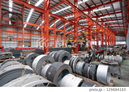 A gargantuan warehouse brims with a labyrinth of gleaming steel beams crisscrossing overhead, creating a metallic canopy. There are steel sheet roll and metal sheets on the floor. 129884183