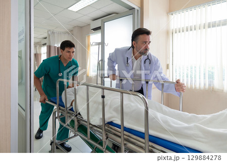 Two healthcare worker are hurriedly transferring a patient on a stretcher in a bright hospital emergency room during the day, illustrating urgency and teamwork in medical care. Two healthcare worker are hurriedly transferring a patient on a stretcher in a bright hospital emergency room during the day, illustrating urgency and teamwork in medical care. 129884278