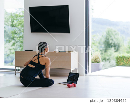 A young woman in sportswear is sitting in the living room and preparing for online training while using a laptop. 129884433