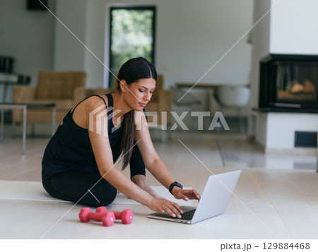 A young woman in sportswear is sitting in the living room and preparing for online training while using a laptop. A young woman in sportswear is sitting in the living room and preparing for online training while using a laptop. 129884468