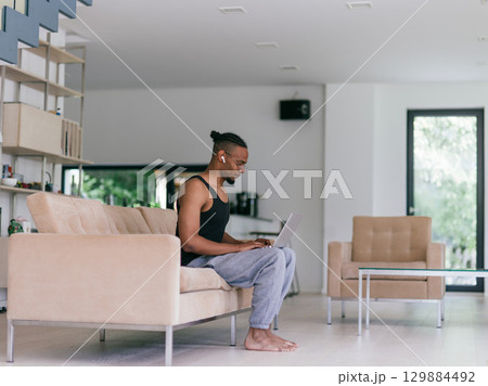 African American man in glasses sitting at a table in a modern living room, using a laptop for business video chat, conversation with friends and entertainment. 129884492