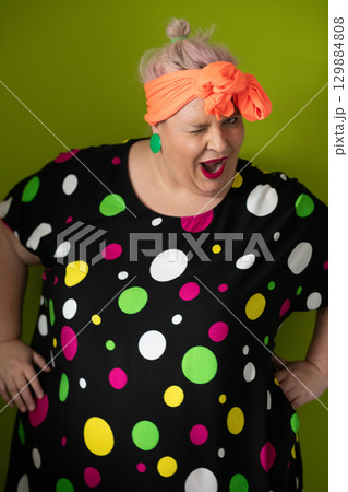 Plus size woman in vibrant polka dot outfit making a comic shrug gesture with outstretched hands and amusing expression. 129884808
