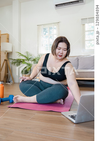 Overweight Woman Exercising at Home with Yoga Mat and Laptop for Online Fitness Class 129884894