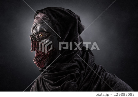 Portrait of a monster with sharp fangs with hooded cape over dark foggy background. Portrait of a monster with sharp fangs with hooded cape over dark foggy background. 129885085