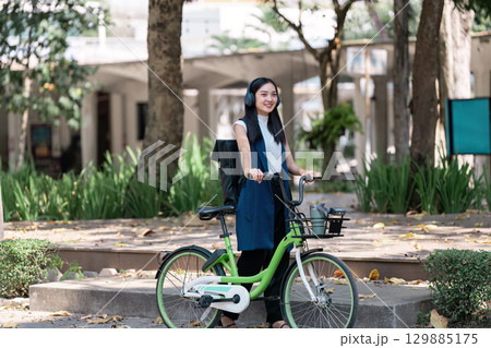 Young Woman Enjoying a Leisurely Ride on a Green Bicycle in a Park Setting with Trees and Modern Architecture in the Background 129885175