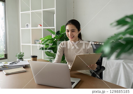 Professional Businesswoman Working Remotely in Modern Office with Laptop and Clipboard, Surrounded by Greenery and Minimalist Decor Professional Businesswoman Working Remotely in Modern Office with Laptop and Clipboard, Surrounded by Greenery and Minimalist Decor 129885185
