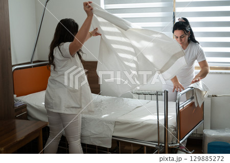 Two nurses in uniforms cleaning and organizing a hospital room by changing the bed linens and ensuring a hygienic environment for patients. 129885272